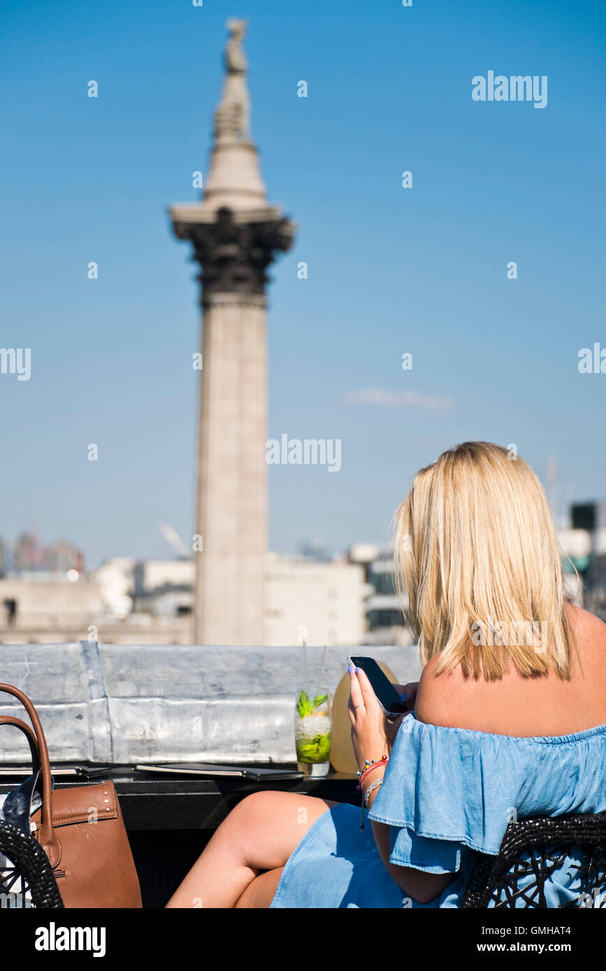 Ritratto verticale di una donna gustando un cocktail affacciato su Nelson's colonna in Trafalgar Square, Londra, sotto il sole. Foto Stock