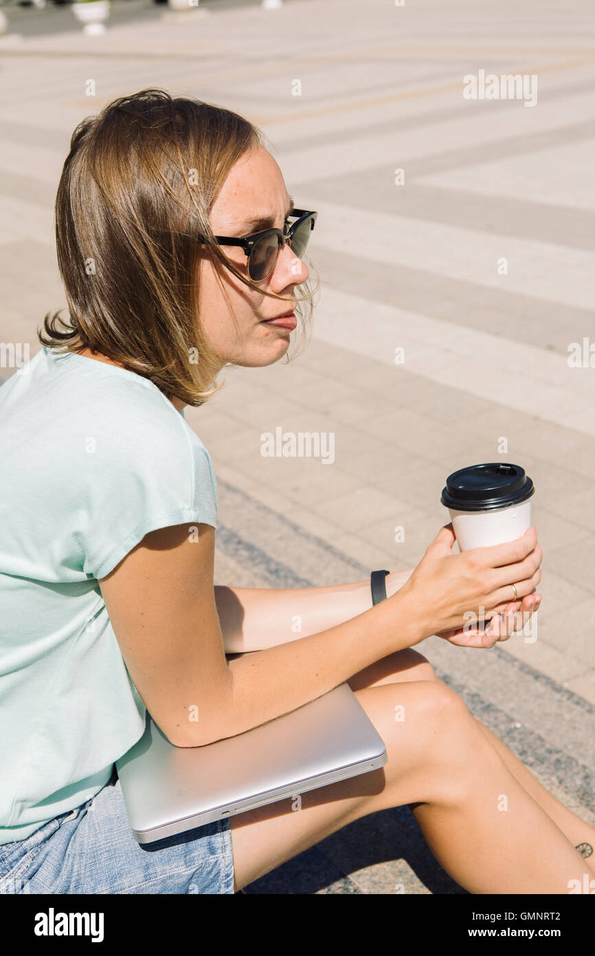 Pensieroso giovane donna con tazza di caffè e il computer portatile in strada Foto Stock