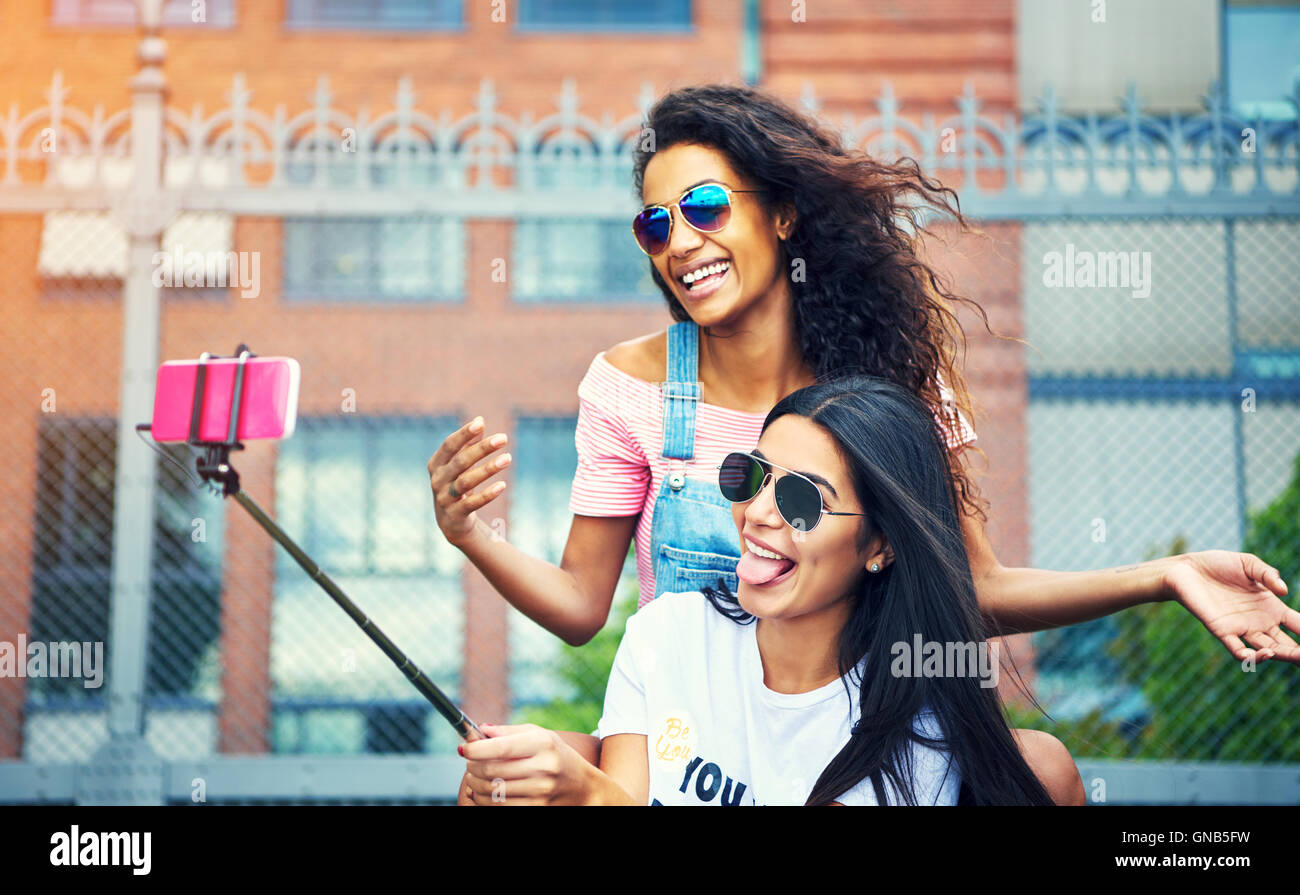 Coppia di amici felice facendo facce buffe tenendo un autoritratto al di fuori con il rosa telefono cellulare attaccato al bastone selfie pole Foto Stock