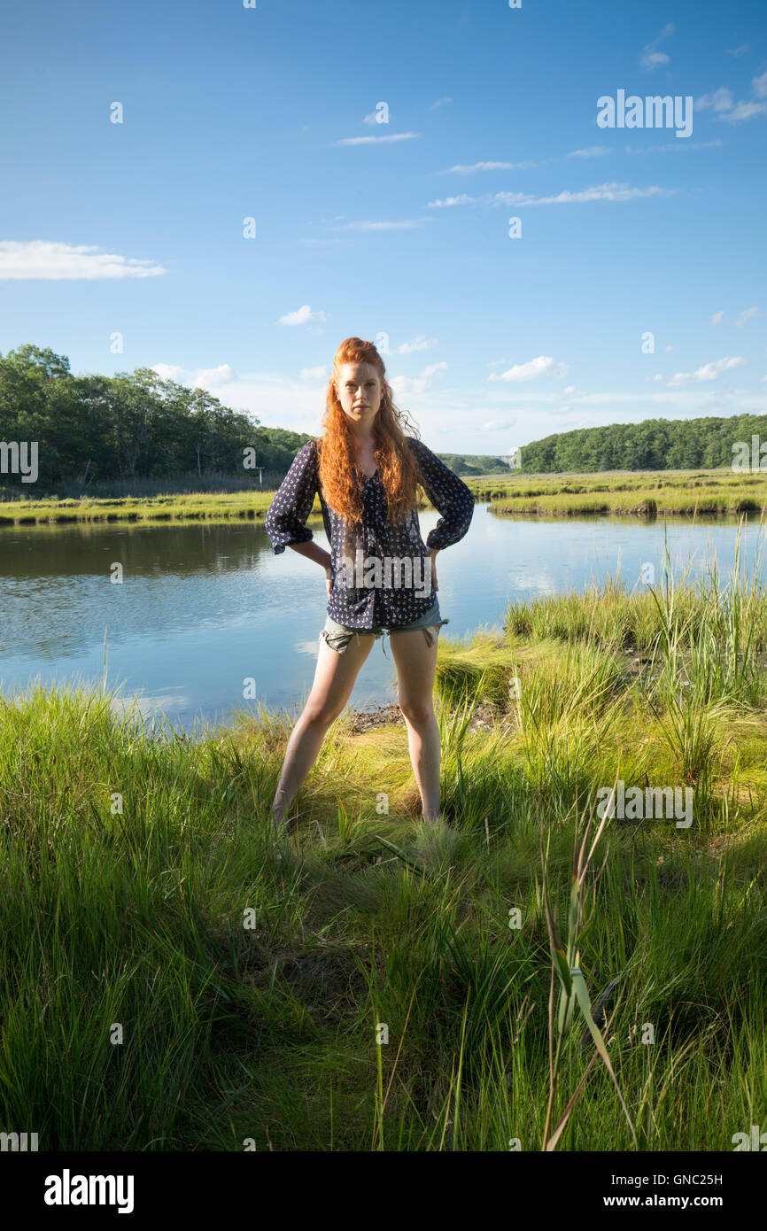 Giovane donna con lunghi capelli rossi permanente sulla guardia in un tranquillo marsh in estate, indossare pantaloncini corti e una maglietta. Foto Stock