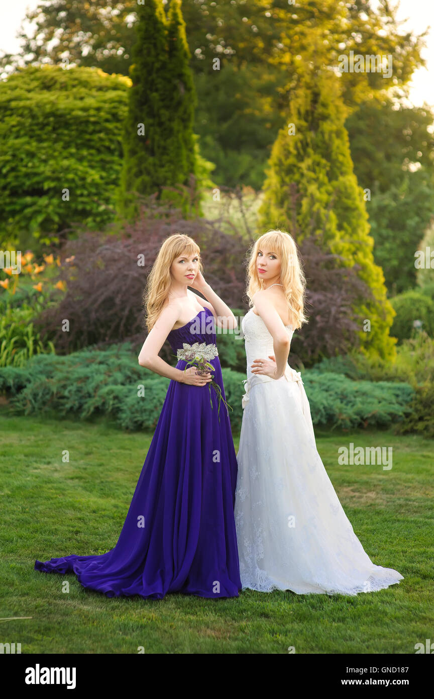 Sposa e damigella al di fuori. Due ragazze in posa sul prato verde. Sposa abiti da sposa. Damigella in un viola abito da sera. Foto Stock