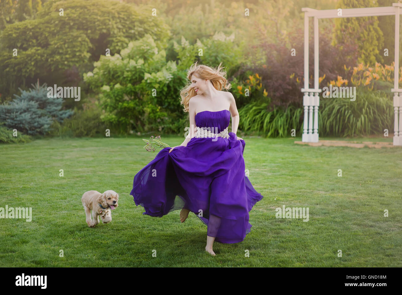 Bella ragazza che corre su un prato con il cane. Damigella in un viola abito da sera. Foto Stock