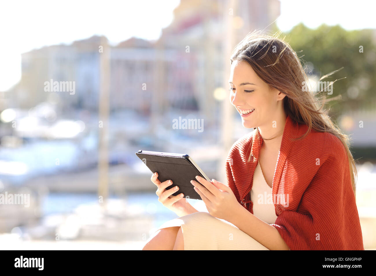 Bella donna seduti all'aperto e utilizzando un tablet a guardare i contenuti multimediali in una soleggiata giornata di vento in un porto della costa Foto Stock