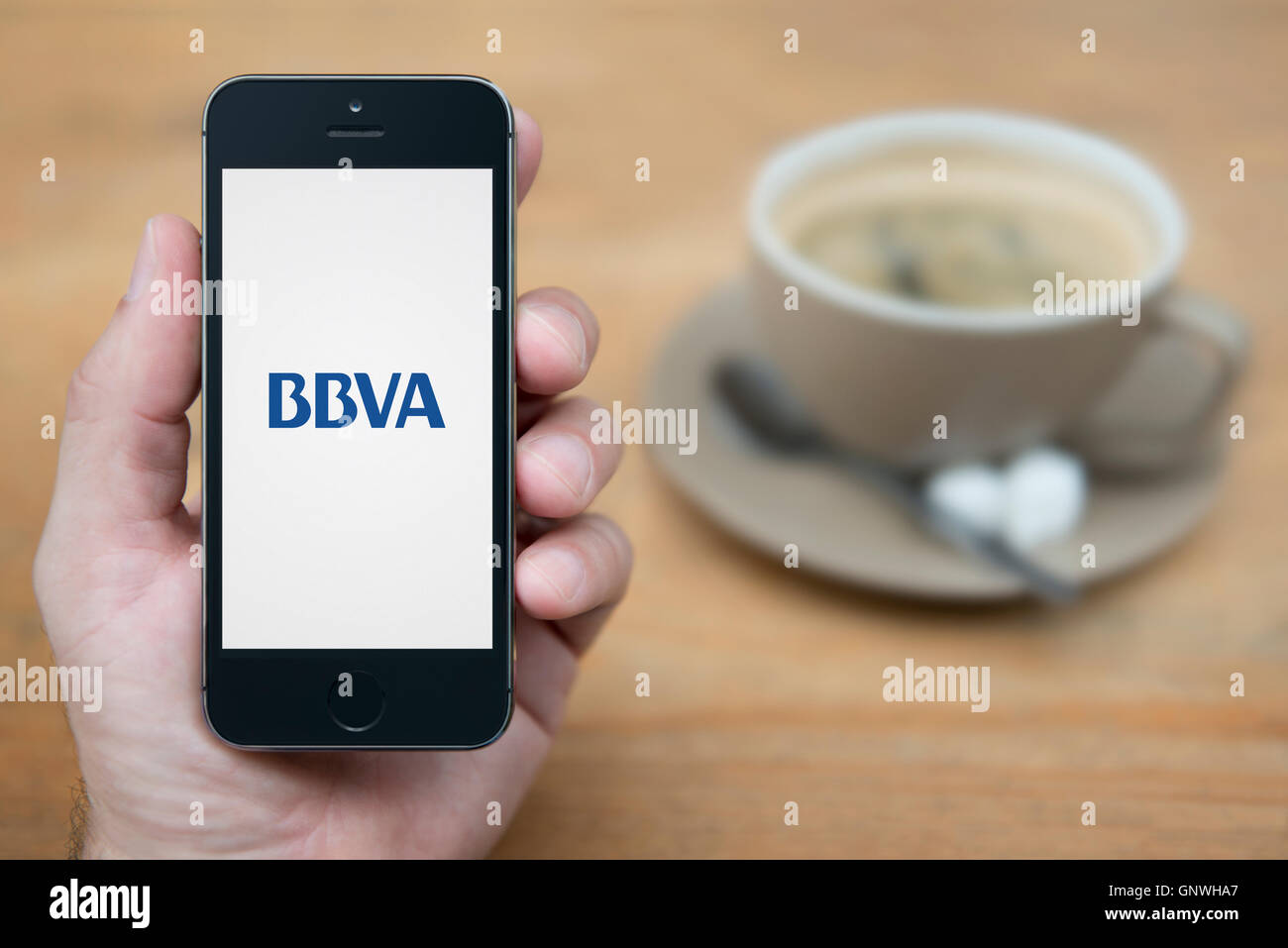 Un uomo guarda al suo iPhone che visualizza il BBVA banca logo, mentre sat con una tazza di caffè (solo uso editoriale). Foto Stock