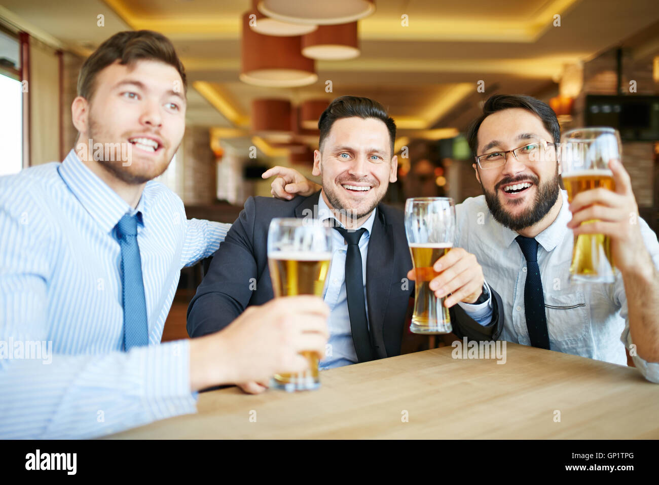 Tre formalmente vestiti uomini d'affari in legami con trasmissione via IR di sorrisi guardando la fotocamera per celebrare il successo con la birra nel bar. Foto Stock