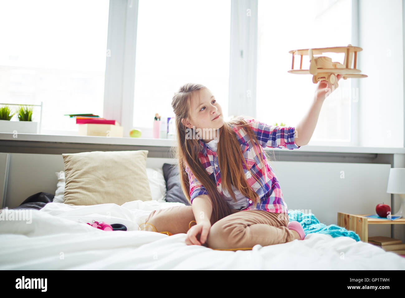 Con i capelli lunghi schoolgirl in rosa Maglietta a scacchi giocare con legno aereo giocattolo sul letto nella sua camera da letto Foto Stock Con i capelli lunghi schoolgirl in rosa Maglietta a scacchi giocare con legno aereo giocattolo sul letto nella sua camera da letto Foto Stock