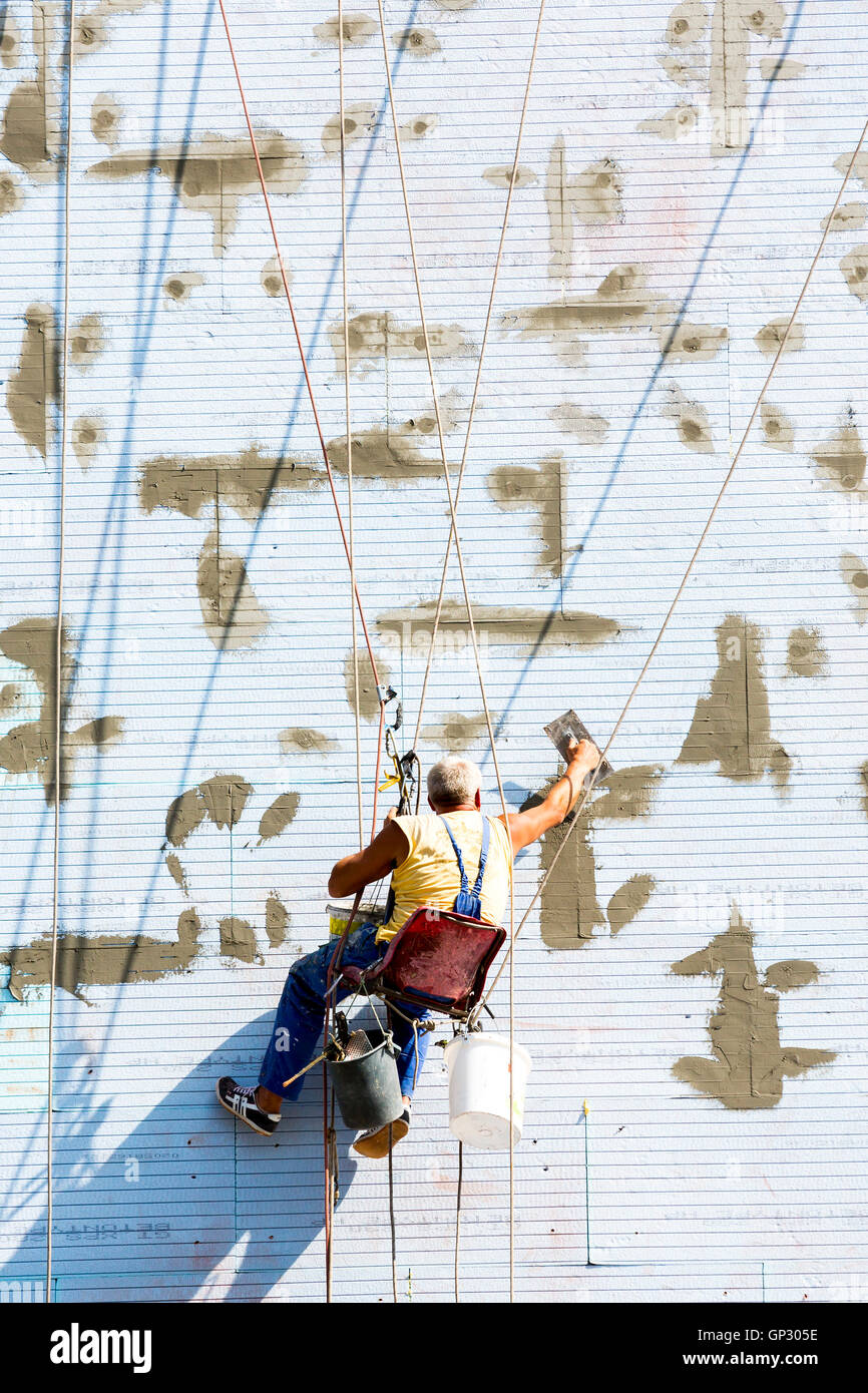 Un lavoratore a mettere su un edificio di termica e isolamento acustico funziona appeso su funi sulla parete dell'edificio. Foto Stock