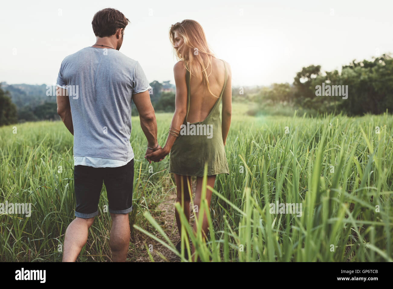 Vista posteriore del tiro del giovane uomo e donna holding hands camminare insieme attraverso il campo di erba. Coppia giovane a piedi attraverso grassy ro Foto Stock
