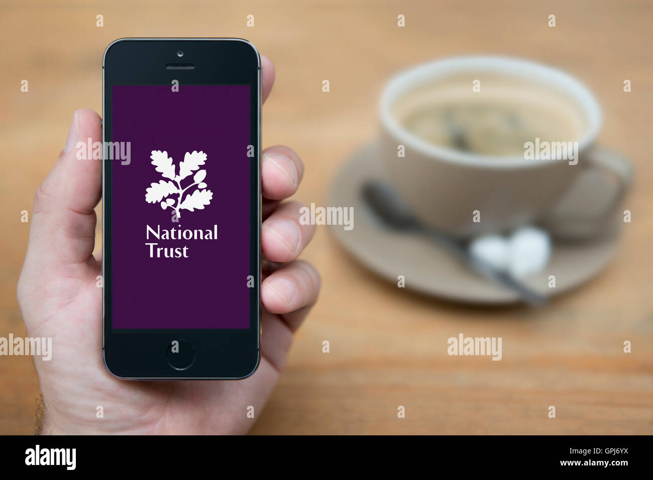 Un uomo guarda al suo iPhone che visualizza il National Trust logo, mentre sat con una tazza di caffè (solo uso editoriale). Foto Stock
