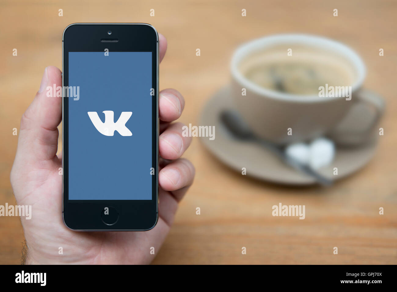 Un uomo guarda al suo iPhone che visualizza il VK russo social media logo, mentre sat con una tazza di caffè (solo uso editoriale). Foto Stock