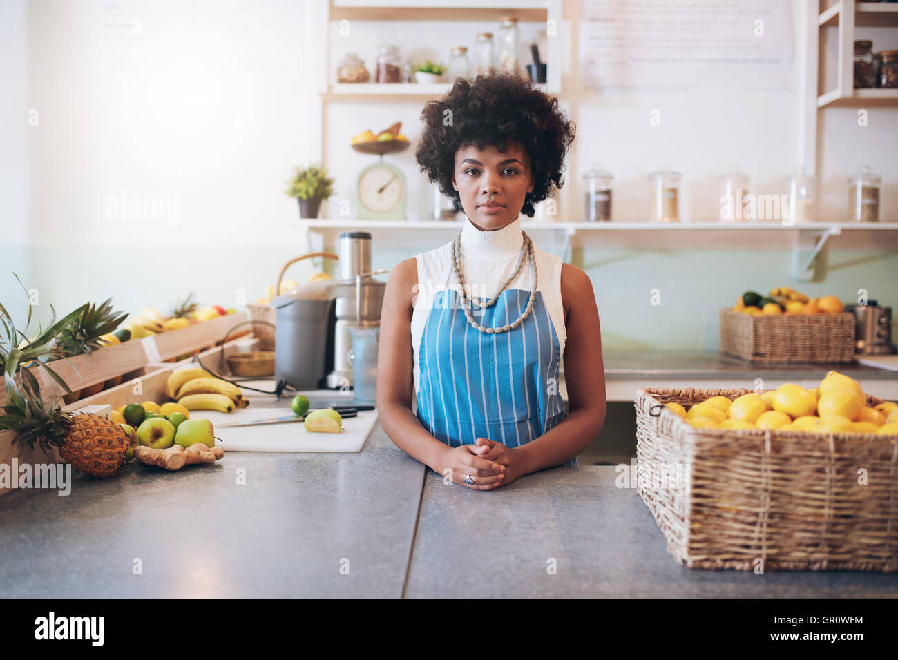 Ritratto di giovane donna africana che indossa il grembiule in piedi dietro il succo di bar counter guardando la fotocamera. Foto Stock Ritratto di giovane donna africana che indossa il grembiule in piedi dietro il succo di bar counter guardando la fotocamera. Foto Stock