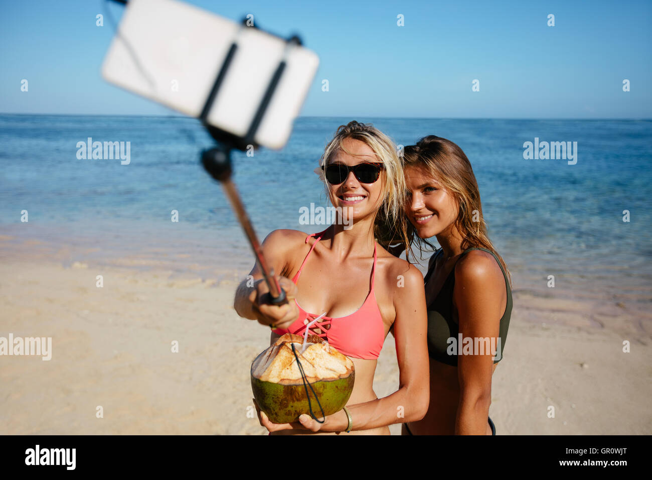 Due giovani donne in costume da bagno sulla spiaggia prendendo autoritratto con smart phone su selfie stick. Foto Stock