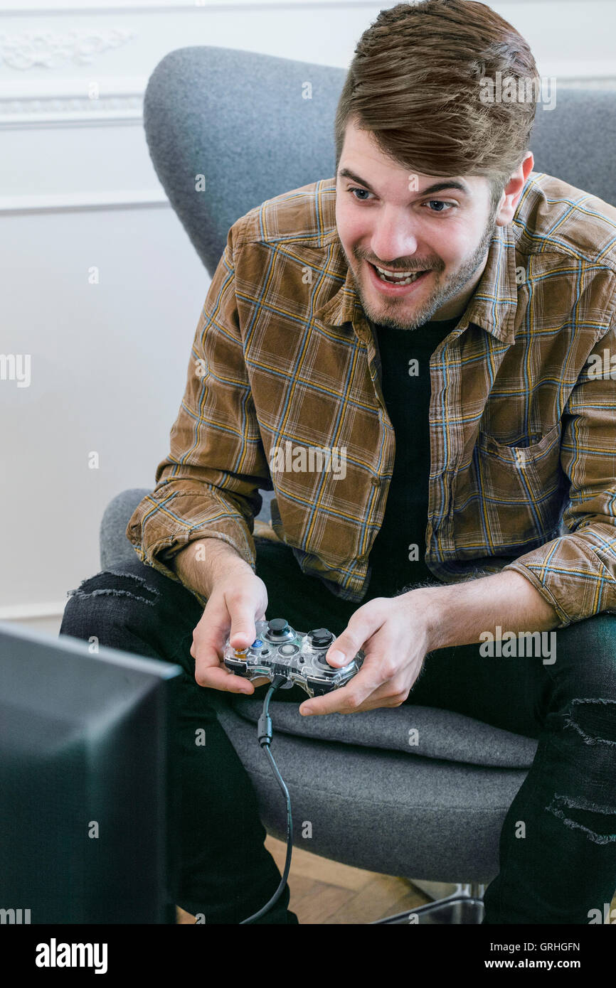 Un giovane dai capelli scuri uomo si inclina in avanti nella sua sedia mentre si tiene un controller di gioco. Foto Stock