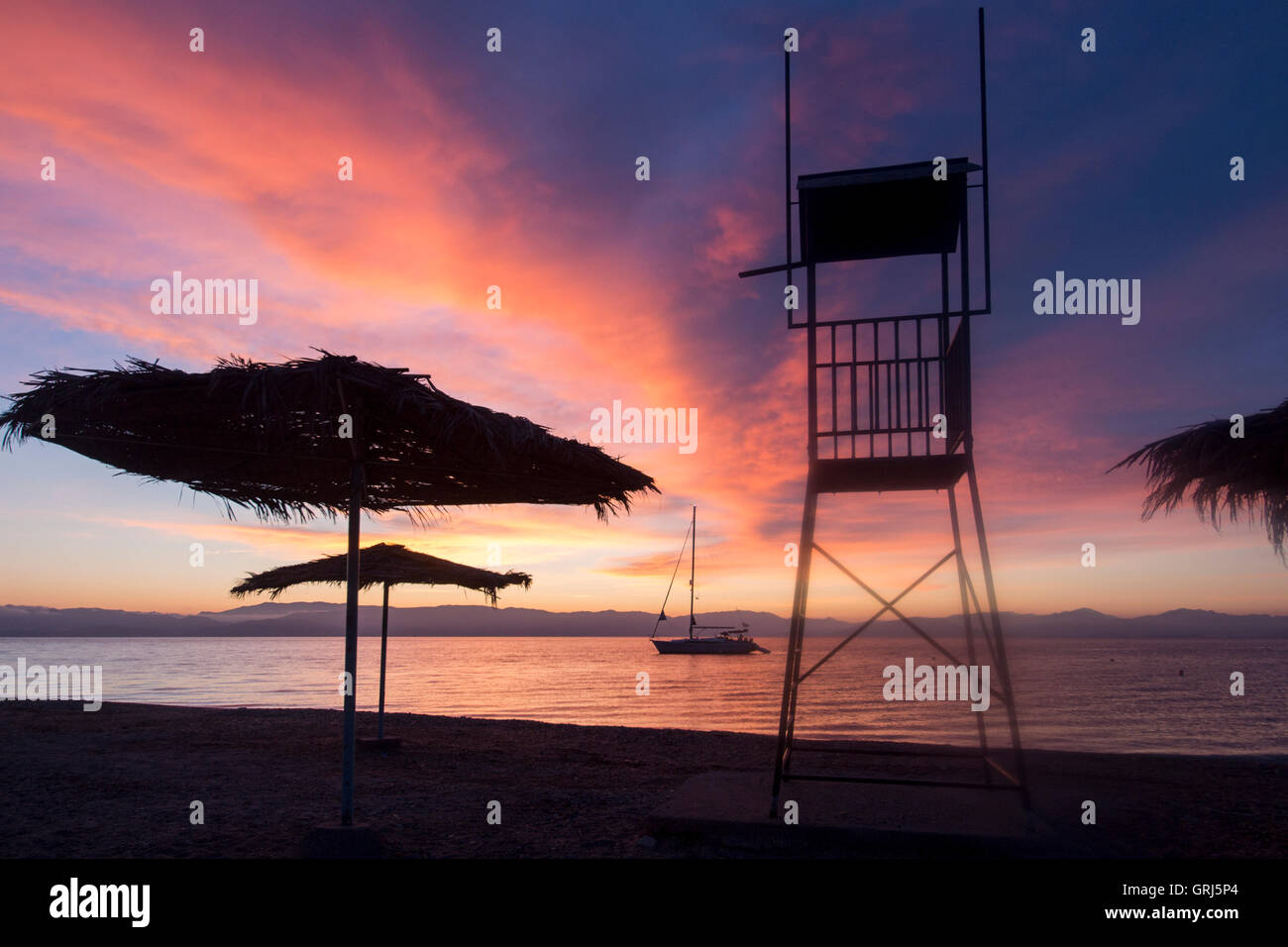 Benitses Grecia sunrise sulla spiaggia con ombrelloni di paglia, barca e torre bagnino stagliano Foto Stock