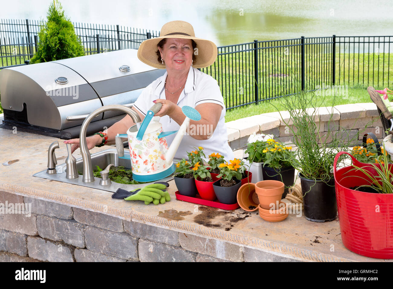 Attraente gentile signora anziana in una vasta-colmato la paglia sun hat innaffiando le sue houseplants su un patio esterno di riempimento del possibile Foto Stock