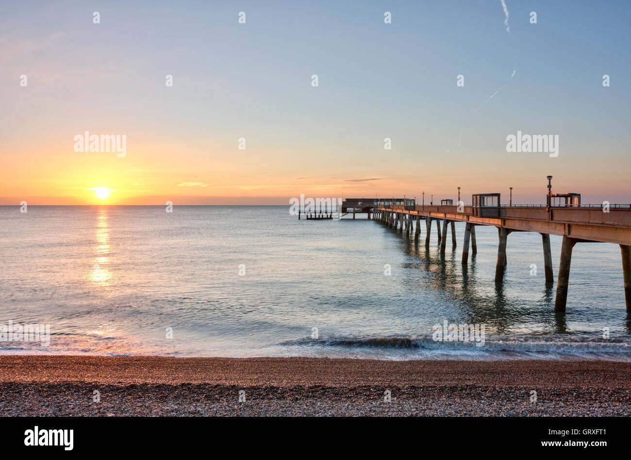 Sunrise, sunup nel cielo blu chiaro con la striscia arancione sul mare di vongole oltre il molo di cemento alla Kent città costiera di trattativa. Spiaggia di ciottoli in primo piano. Foto Stock