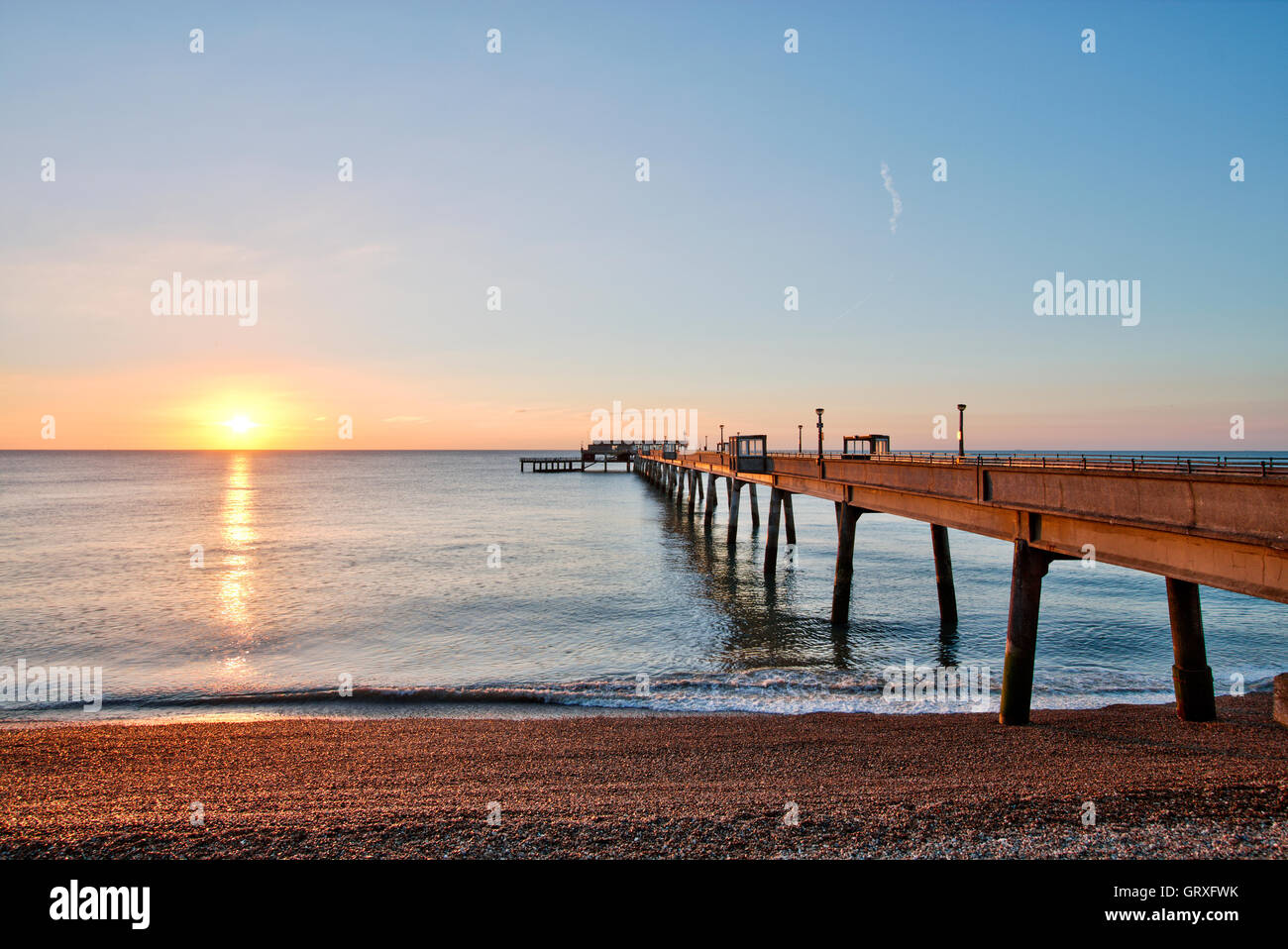 Sunrise, sunup nel cielo blu chiaro con la striscia arancione sul mare di vongole oltre il molo di cemento alla Kent città costiera di trattativa. Spiaggia di ciottoli in primo piano. Foto Stock