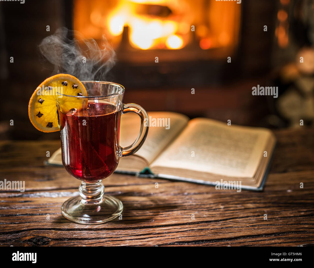 Vin brulé e un libro sul tavolo di legno. Camino con fuoco caldo sullo sfondo. Foto Stock