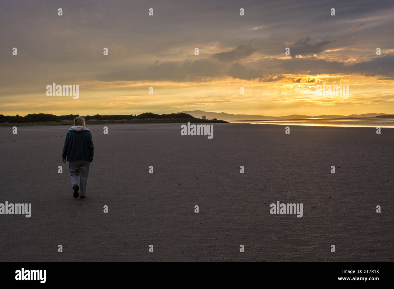 Una donna sola camminando sul Solway Firth spiaggia al tramonto, Bowness-on Solway, Cumbria, Inghilterra. Resti di giunzione Solway viadotto ferroviario Foto Stock