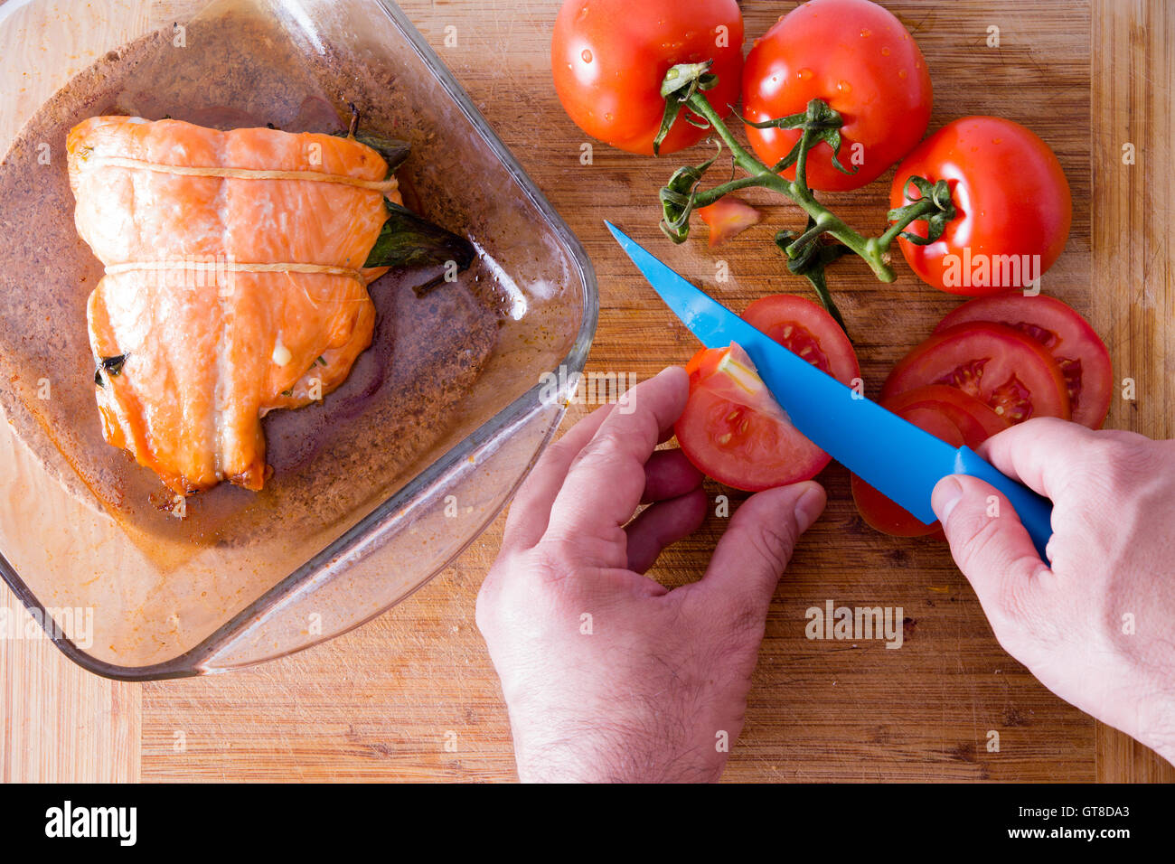 Vista ravvicinata al di sopra delle mani di un maschio di chef per affettare i pomodori con un coltello blu per accompagnare un forno a bistecca di salmone Foto Stock