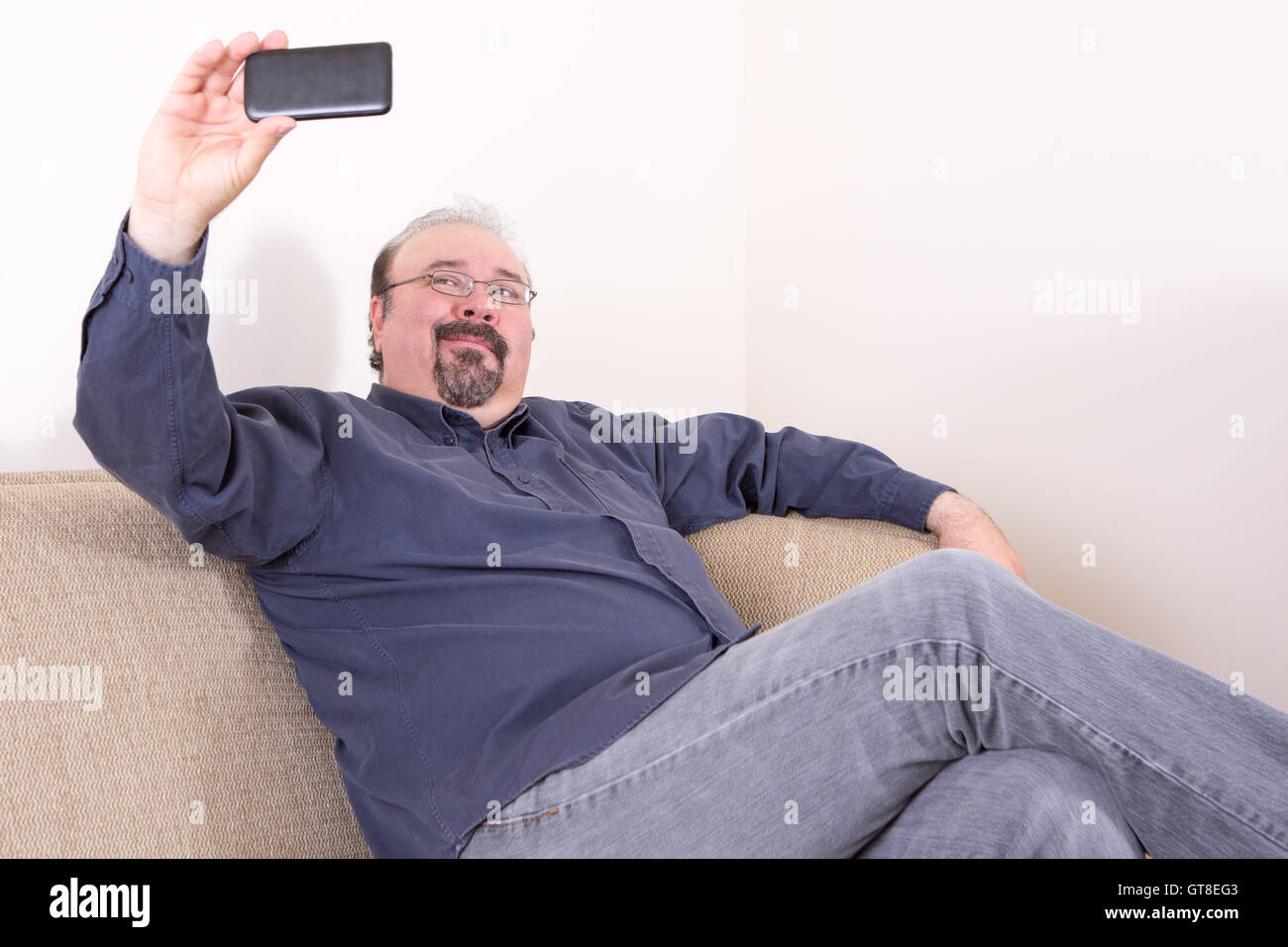 Di mezza età portly uomo che indossa casual dark maglietta blu e grigio jeans mentre è seduto sul divano e tenendo selfie porta orizzontale Foto Stock
