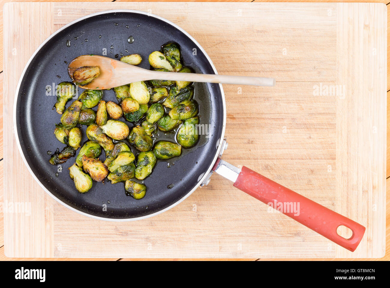 Top down prospettiva in prima persona di deliziosi i cavoletti di Bruxelles cotto in padella antiaderente con il cucchiaio di legno oltre il bordo di taglio Foto Stock