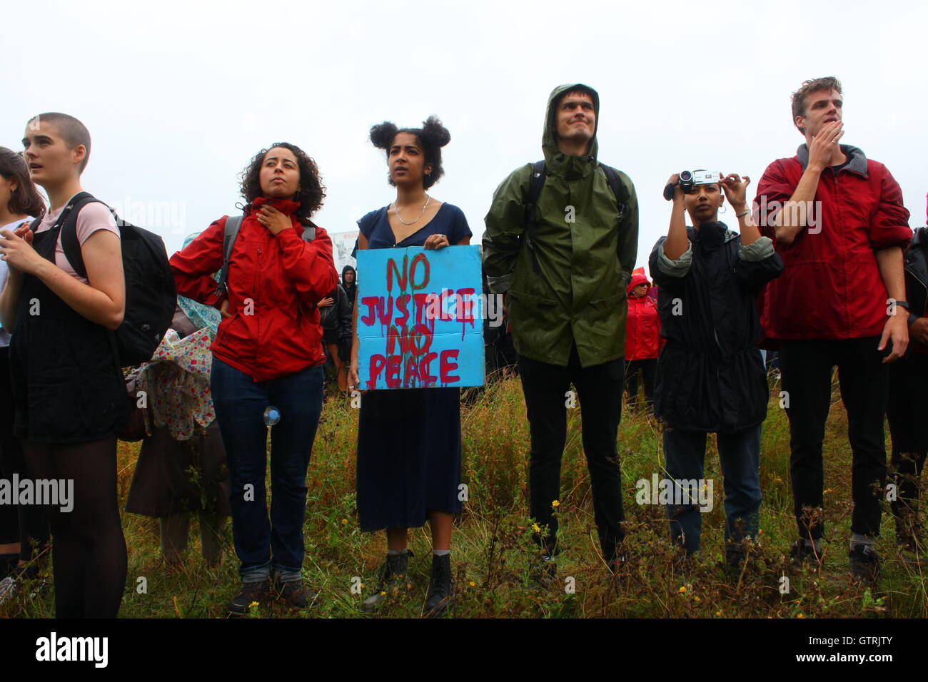 Milton Ernest, Bedford, Regno Unito. 10 settembre 2016. Un gruppo di persone partecipa a una protesta pacifica all'aperto in un'area erbosa, vestita per il tempo umido. Un manifestante ha un cartello fatto a mano con la scritta "NO JUSTICE NO PEACE", mentre un altro documenta l'evento con una telecamera. L'immagine cattura un momento di azione collettiva e di impegno sociale, riflettendo temi di giustizia, attivismo ed espressione pubblica nel Regno Unito. Centinaia di manifestanti protestano contro il recinto del centro di detenzione Yarls Wood chiedendo la chiusura della struttura. Penelope Barritt/Alamy Live News Foto Stock