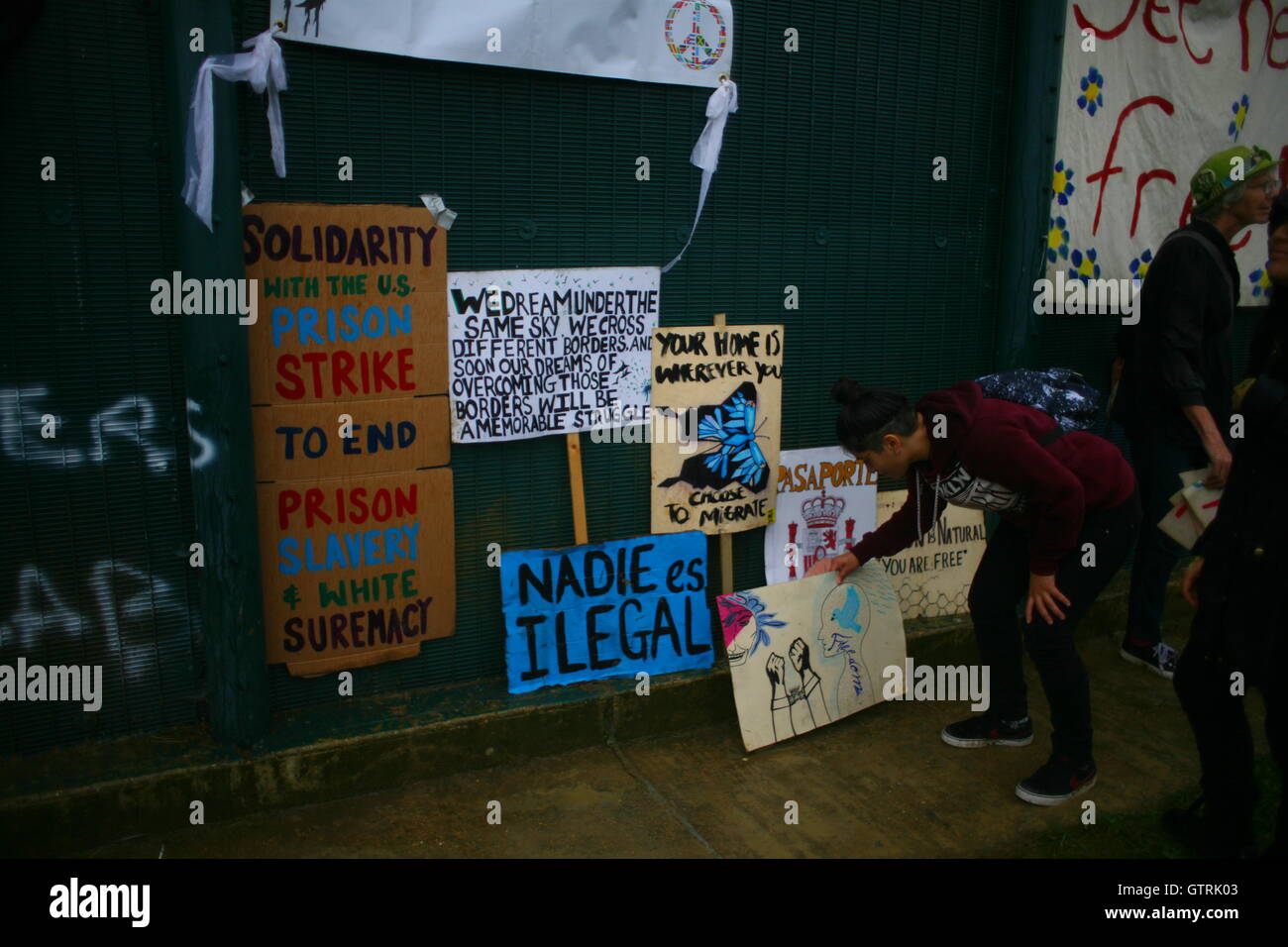 Milton Ernest, Bedford, Regno Unito. 10 settembre 2016. Una collezione di cartelli di protesta esposti contro un muro verde, con persone presenti. I messaggi includono la solidarietà con lo sciopero carcerario americano, l'opposizione alla supremazia bianca, le richieste di attivismo multirazziale e il sostegno ai diritti degli immigrati. I CARTELLI RECITANO “SOLIDARIETÀ CON LO SCIOPERO CARCERARIO AMERICANO PER PORRE FINE ALLA SCHIAVITÙ CARCERARIA E ALLA SUPREMAZIA BIANCA”, “NADIE ES ILEGAL” E “PRIGIONI USA = SCHIAVITÙ MODERNA”. Centinaia di manifestanti protestano contro il recinto del centro di detenzione Yarls Wood chiedendo la chiusura della struttura. Penelope Barritt/Alamy Live News Foto Stock