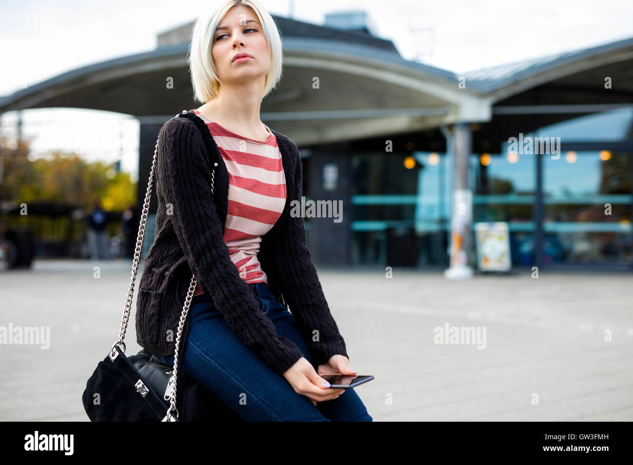 Annoiato donna con telefono cellulare seduti sui bagagli all'esterno della stazione Foto Stock