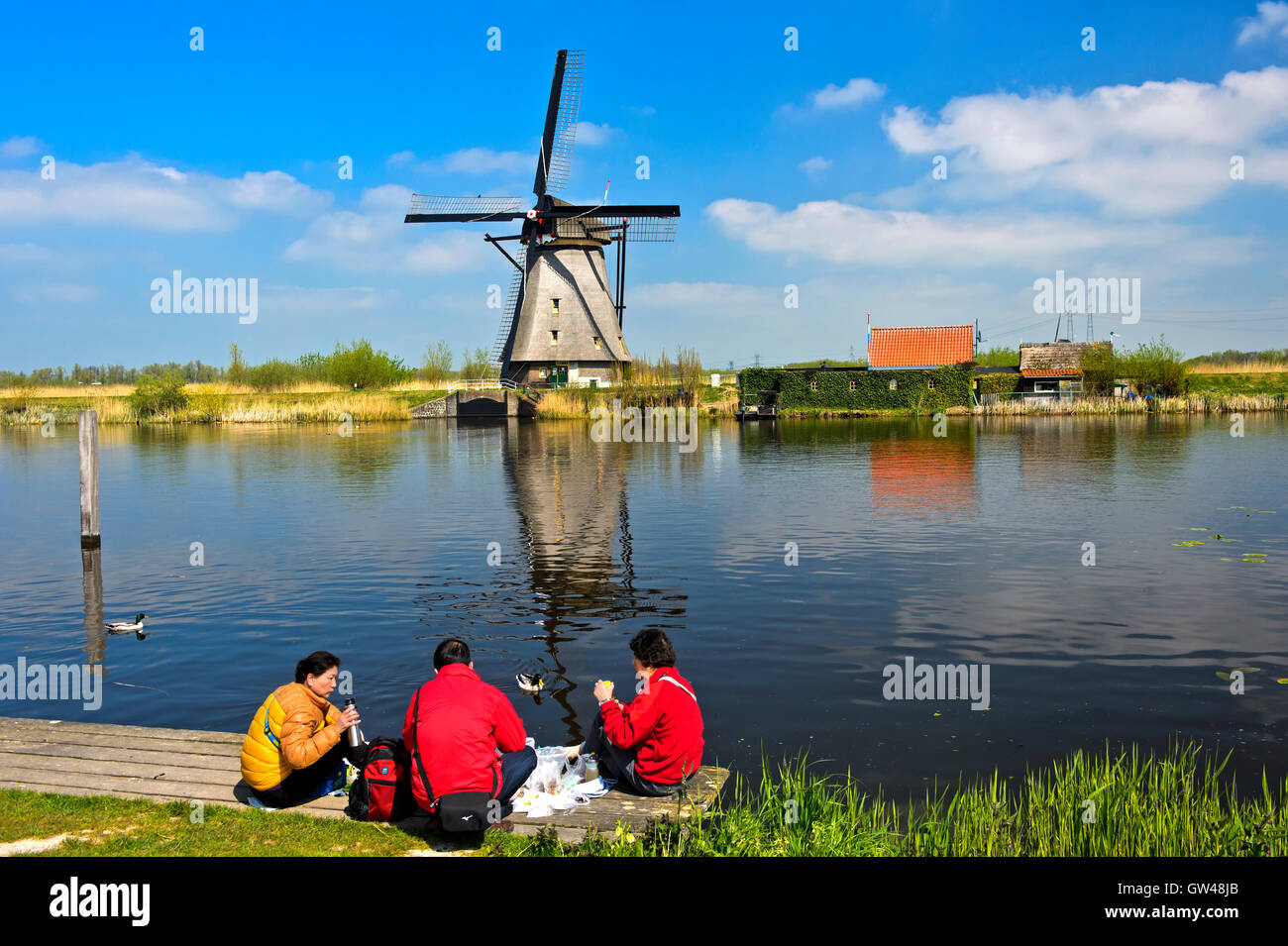 Tre turisti cinesi picnicing a canal con un mulino a vento olandese dietro, Kinderdijk, Alblasserwaard polder, Paesi Bassi Foto Stock