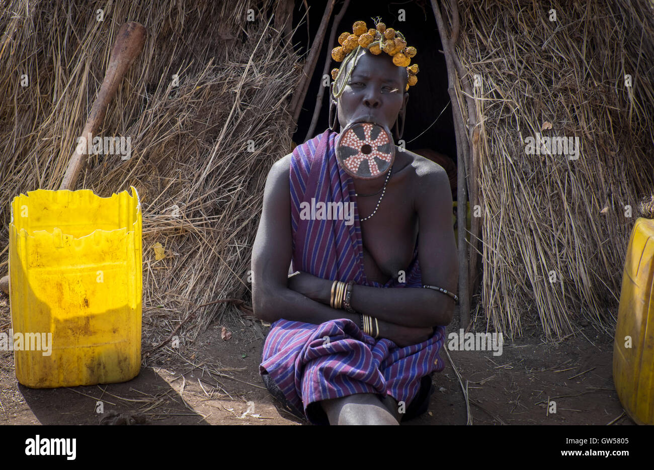 Mursi donna della valle dell'Omo in Etiopia si siede di fronte alla sua capanna con il tetto di paglia con una bocca piastra inserita nel suo labbro inferiore. Foto Stock