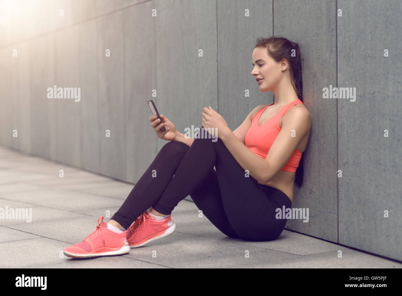 Montare sportivo attraente giovane donna seduta su un cortile lastricato di un edificio commerciale contro un muro grigio controllo del suo cellulare Foto Stock