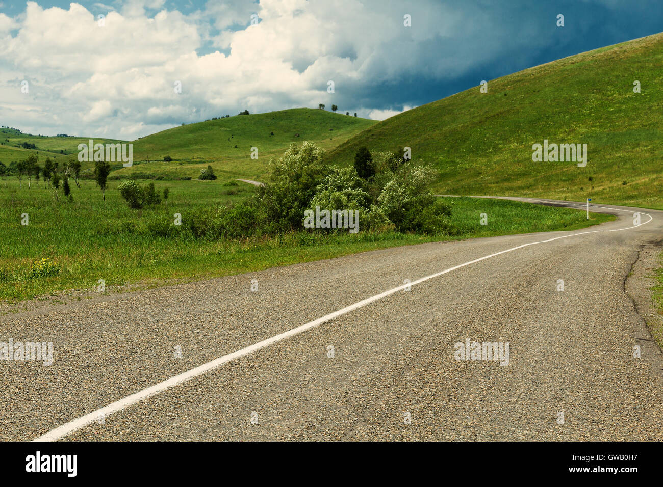 Curve su strada asfaltata con marcatura circondata da verdi colline da estate con tuoni nuvole come sfondo Foto Stock