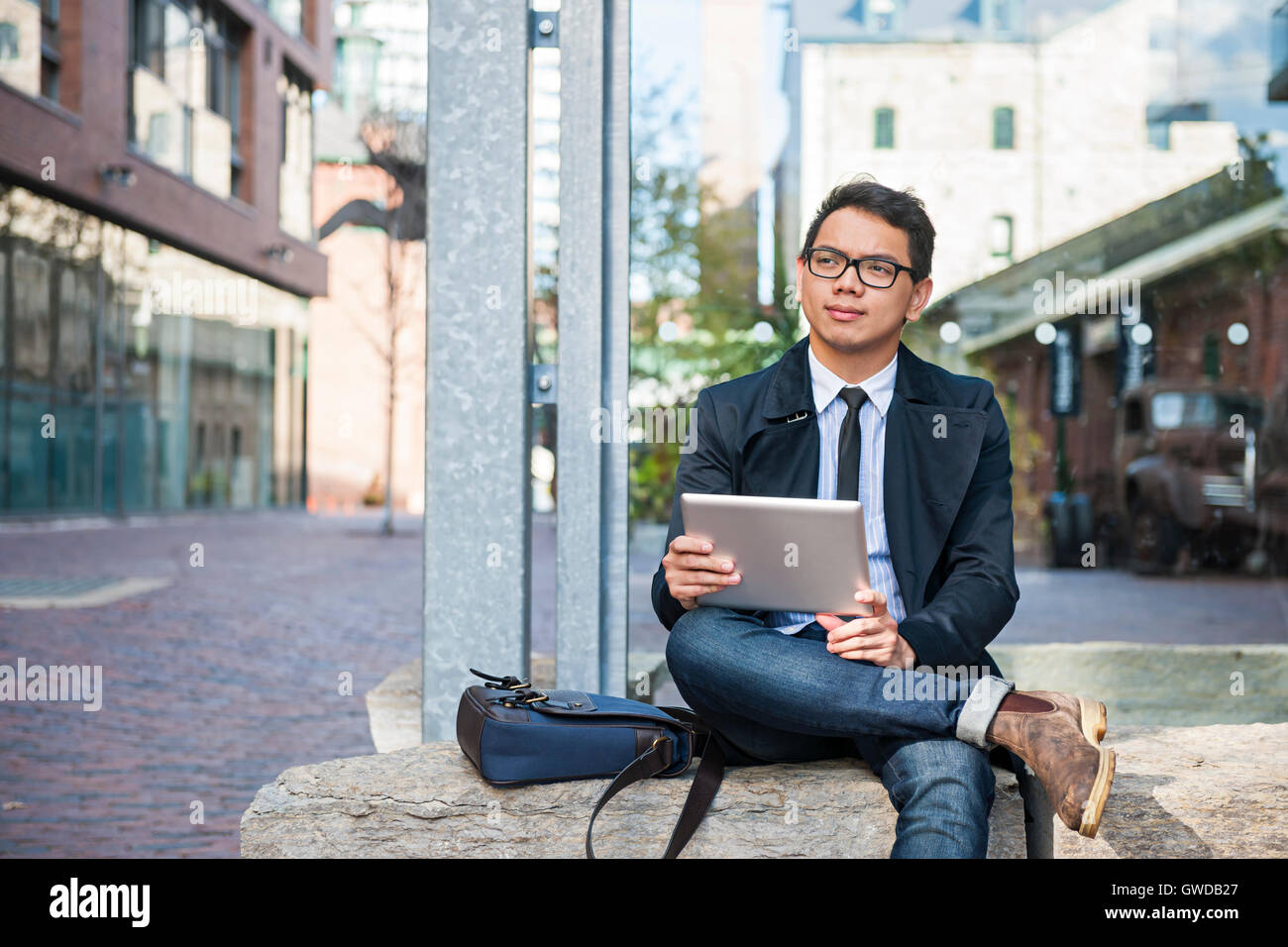 Giovani asiatici business man tenere una tavoletta digitale al di fuori seduta su una strada di città alla ricerca attenta e interessata Foto Stock