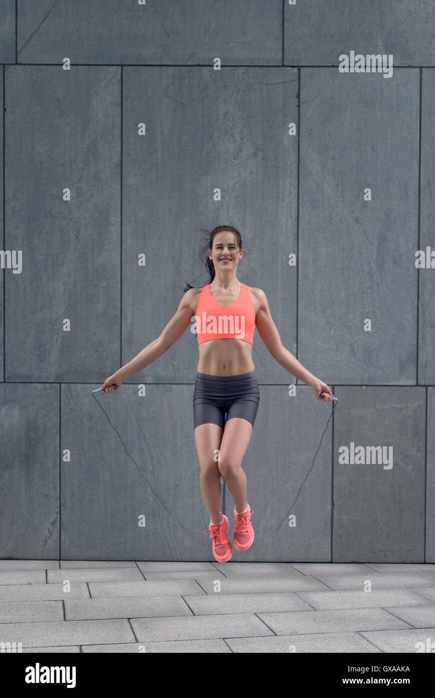 Montare sorridente athletic giovane donna in pantaloncini corti e top salto con la corda al di fuori oltre il blocco di pietra terreno Foto Stock