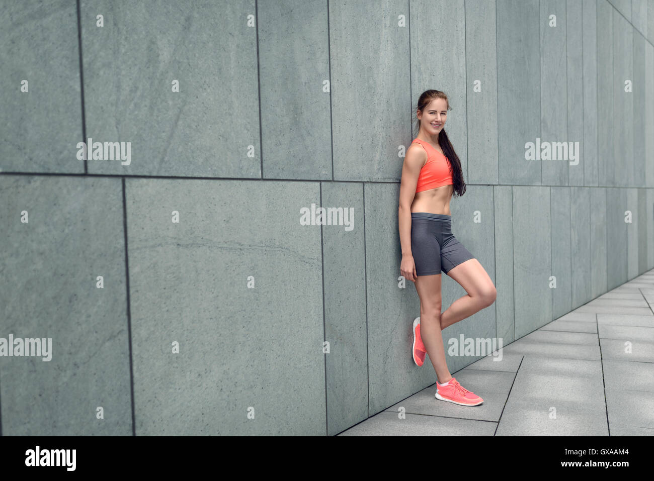 Sportivo attraente giovane donna appoggiata su un grigio muro piastrellato di un edificio urbano con la sua mano per i suoi lunghi capelli castani e un lovel Foto Stock