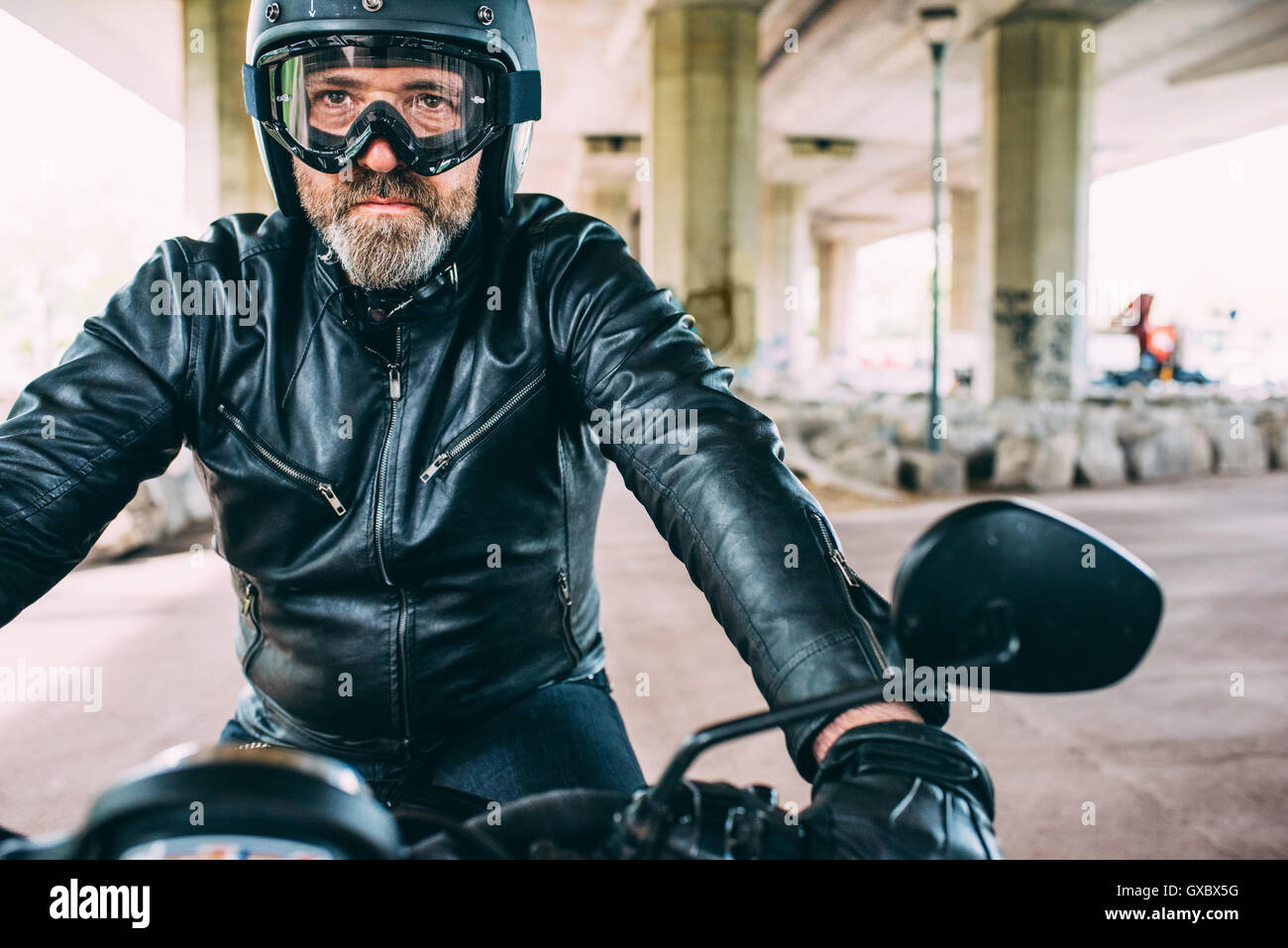 Maschio maturo motociclista che indossa il casco nero e gli occhiali seduto sul motociclo sotto il cavalcavia Foto Stock