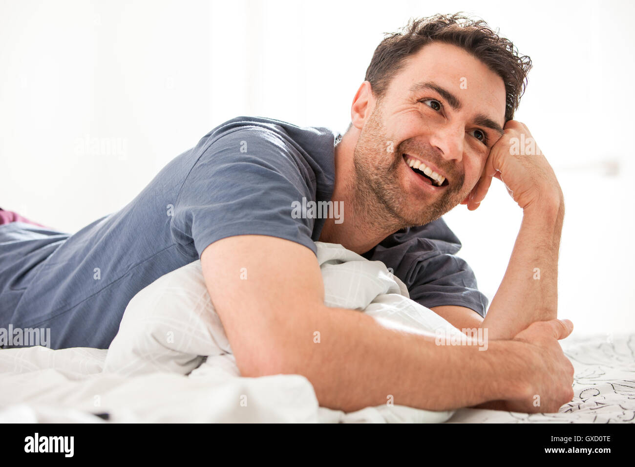 Uomo disteso sul letto, in appoggio sul gomito guardando lontano sorridente Foto Stock