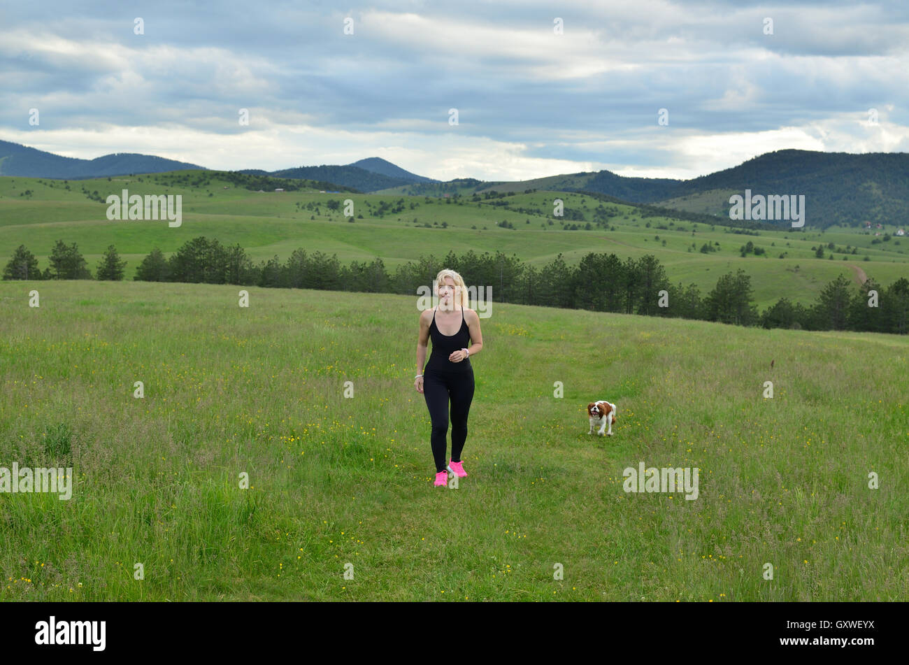 Donna jogging in natura sul giorno nuvoloso mentre il suo cane è in esecuzione accanto a lei Foto Stock