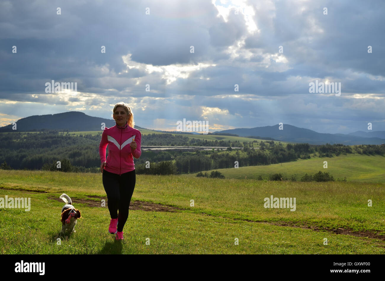 La donna in esecuzione con il suo cane su un prato di montagna al di sotto di cielo nuvoloso in primavera Foto Stock