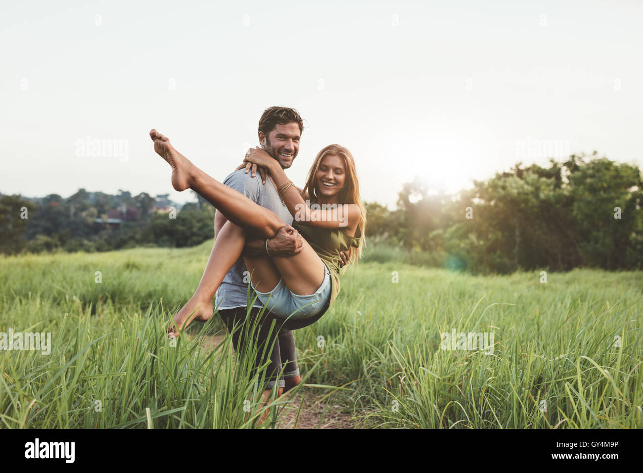 Colpo di giovane portando la sua fidanzata attraverso il campo di erba. Romantico coppia giovane divertendosi in natura in un giorno di estate. Foto Stock