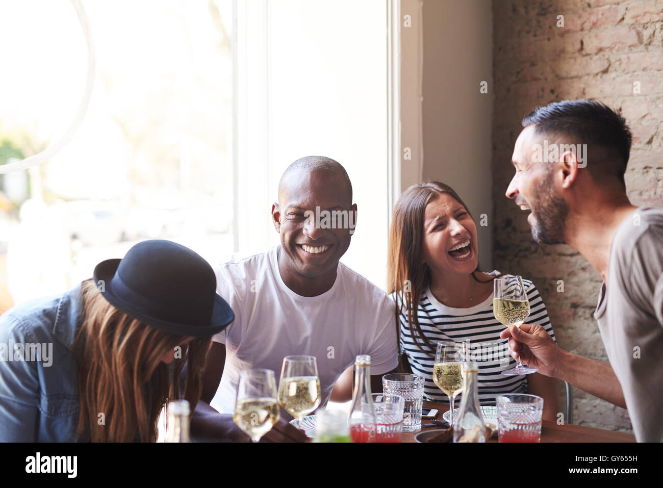 Allegro gruppo di giovani adulti bevuto vino da ridere mentre si è seduti al tavolo per la cena con la luce della finestra e il muro di mattoni in backgrou Foto Stock