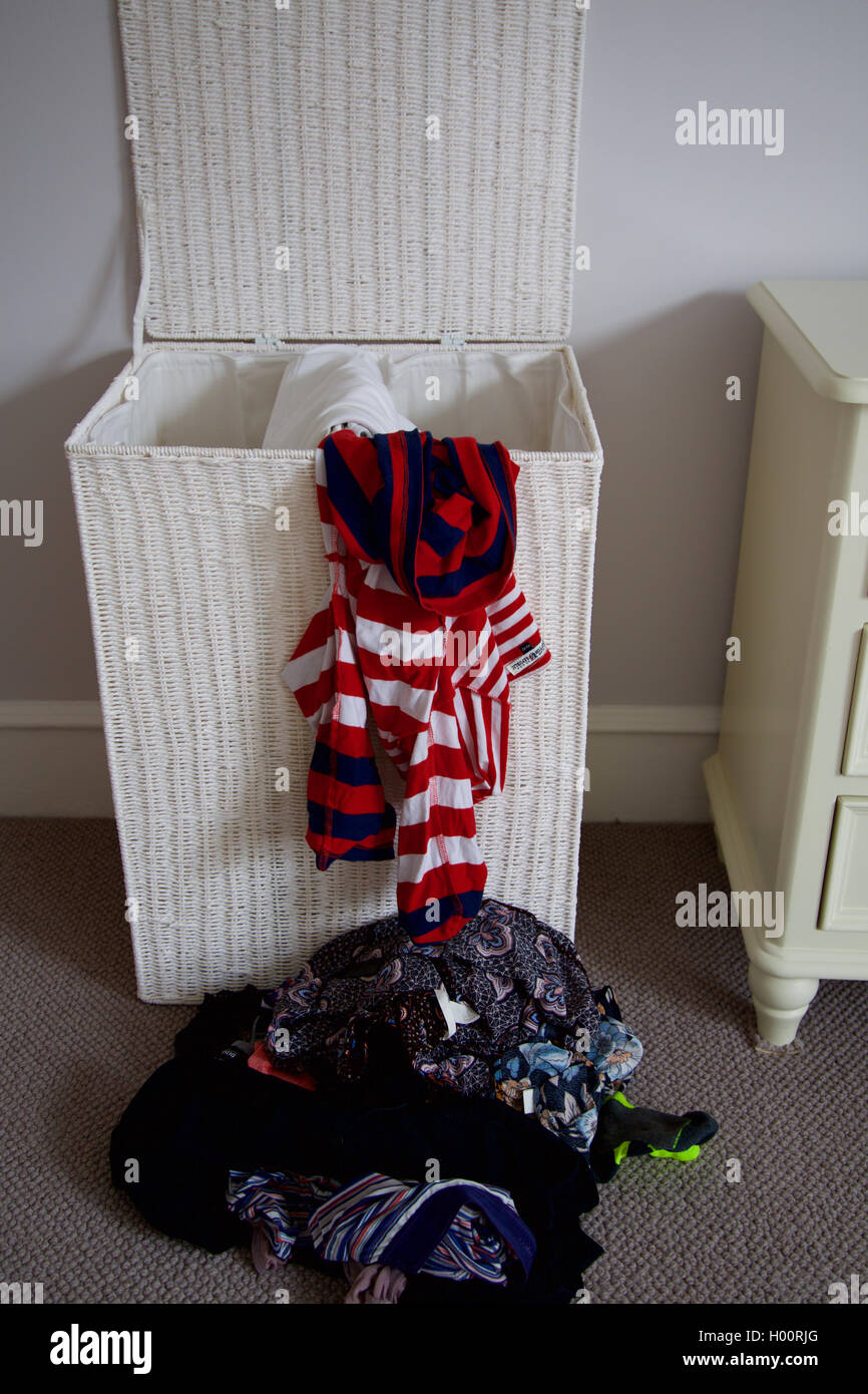 Un coperchio aperto bianco vimini cesto per la biancheria con la pila di roba da lavare vestiti riversandosi sul pavimento di moquette in camera da letto in casa Foto Stock