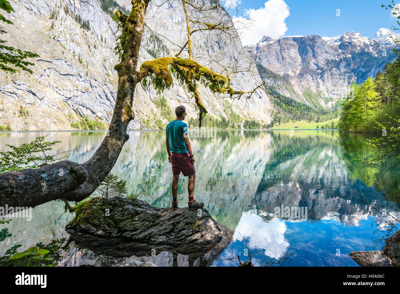 Giovane uomo in piedi sulla pietra in acqua, cercando in distanza, riflesso nel lago Obersee, Salet am Königssee Foto Stock