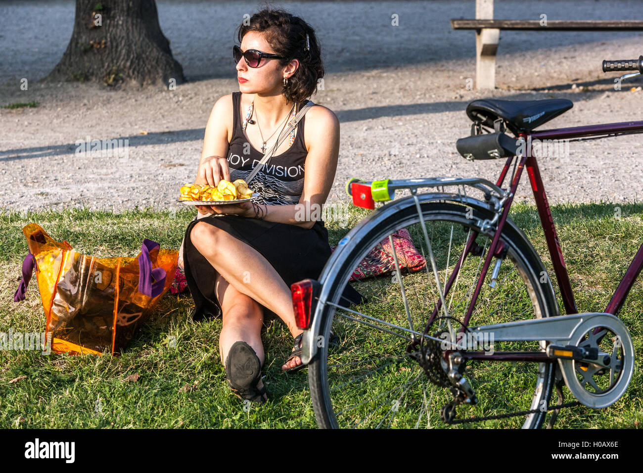 Giovane donna in una canotta della Joy Division che mangia spuntini mentre siede sull'erba accanto alla sua bicicletta nel parco Augarten, Vienna, Austria. Foto Stock