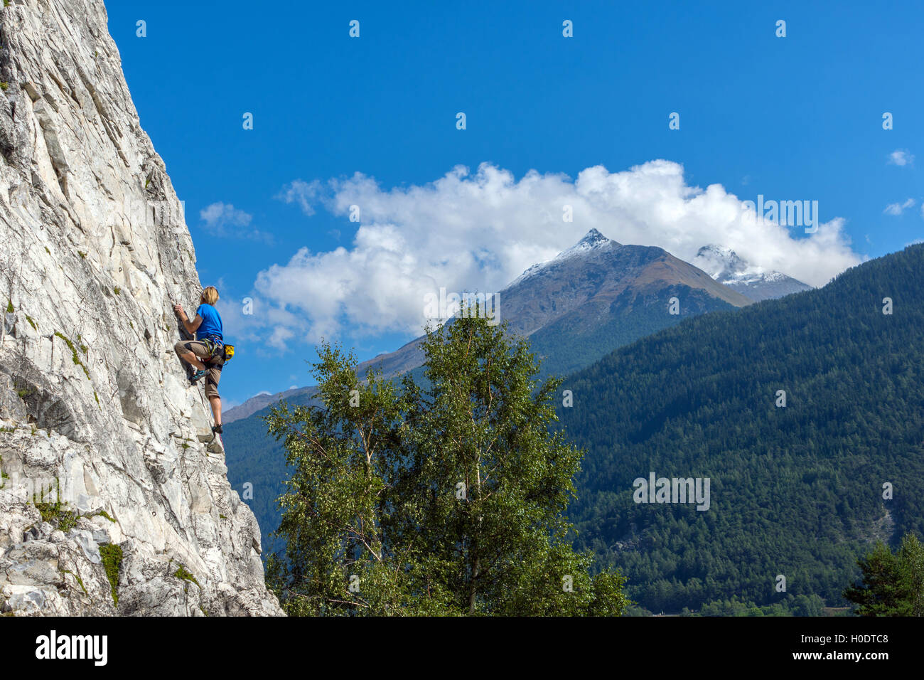 Maschio Slim rocciatore in blu sulla ripida parete di roccia, con cielo blu delle montagne e nuvole Foto Stock