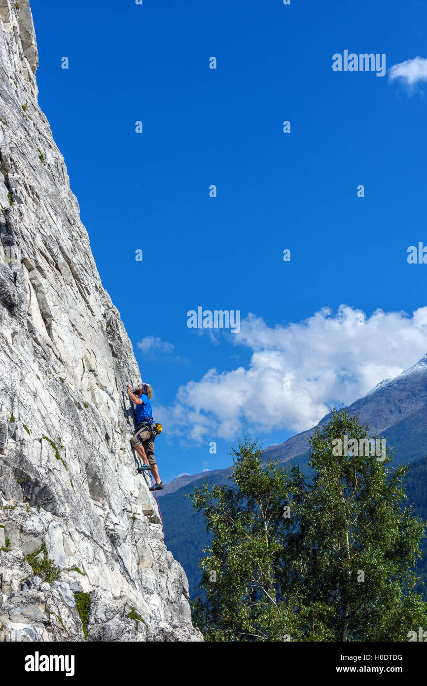Maschio Slim rocciatore in blu sulla ripida parete di roccia, con cielo blu delle montagne e nuvole Foto Stock