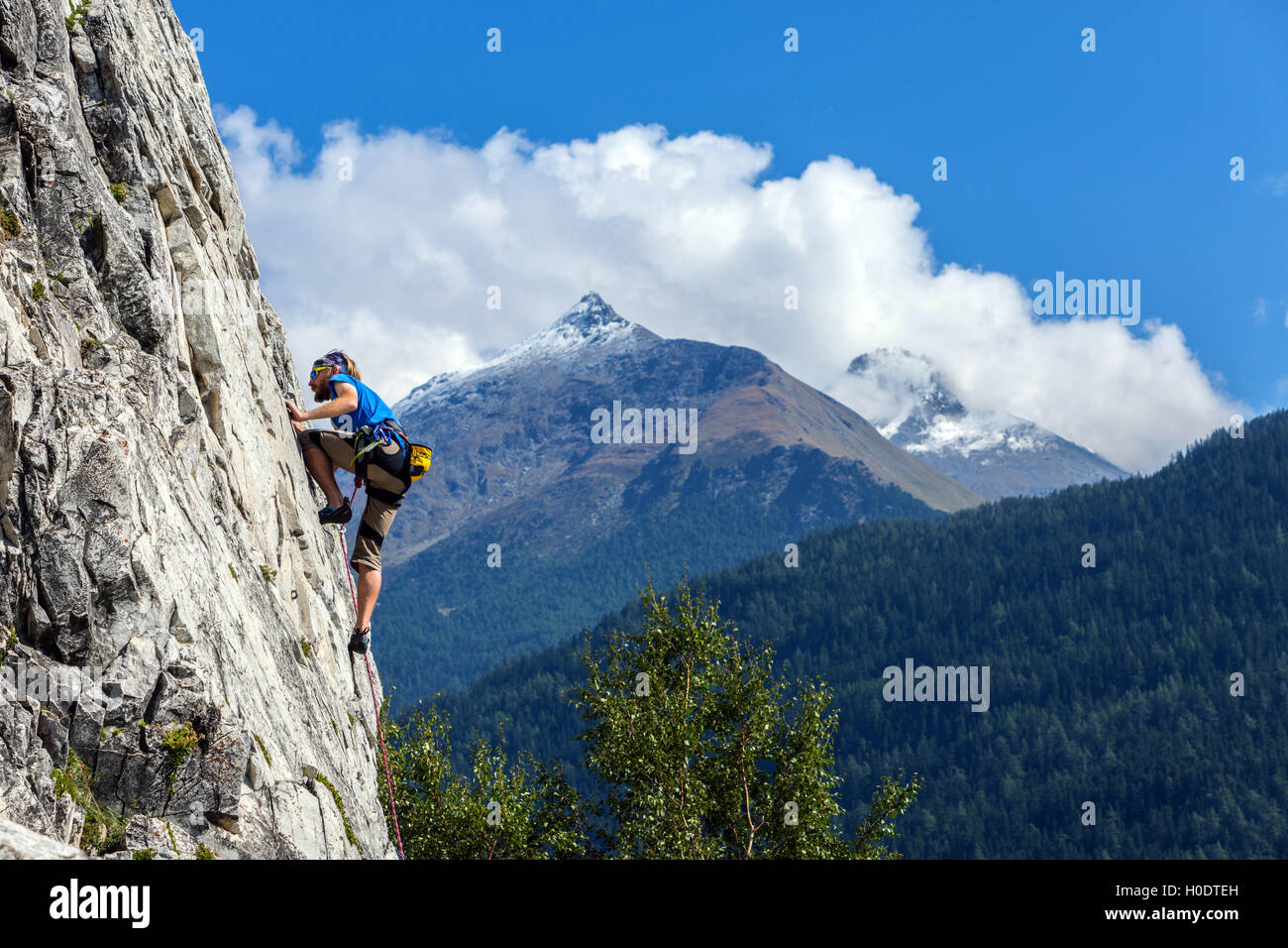 Maschio Slim rocciatore in blu sulla ripida parete di roccia, con cielo blu delle montagne e nuvole Foto Stock