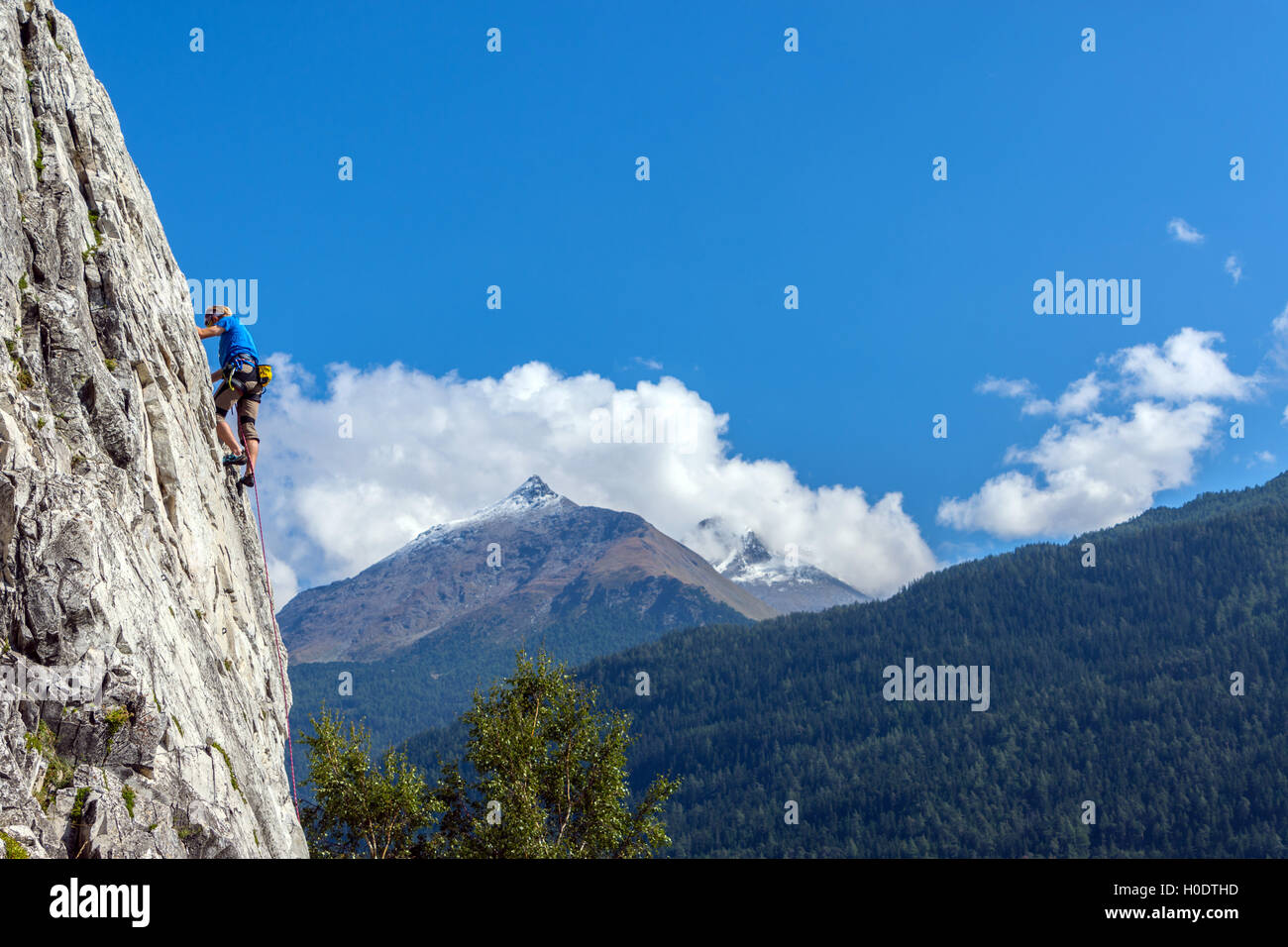 Maschio Slim rocciatore in blu sulla ripida parete di roccia, con cielo blu delle montagne e nuvole Foto Stock