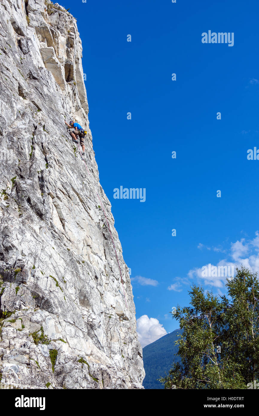 Maschio Slim rocciatore in blu sulla ripida parete di roccia, con cielo blu delle montagne e nuvole Foto Stock